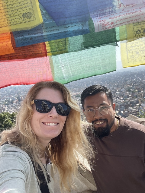       Two people smiling with a cityscape and prayer flags in the background.
  