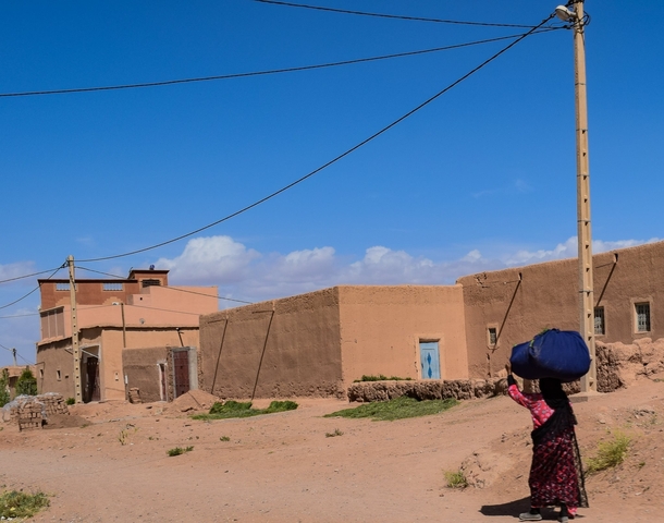       A person carrying a large bundle in a rural setting with mud-brick buildings.
  