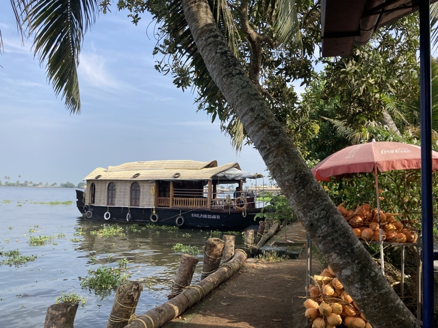 Houseboat on a lake with trees and a coconut stand on the shore.