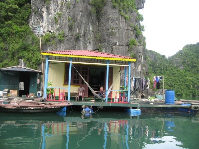 Colorful house on a raft with steep cliffs behind, someone sitting on the porch.