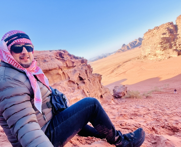       Person sitting on rocky orange sand dunes with rocky formations in the background.
  