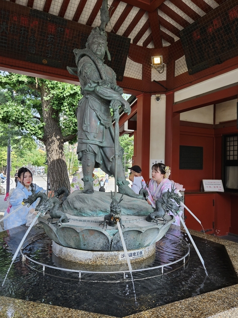 Two women in kimonos posing next to a statue surrounded by trees.