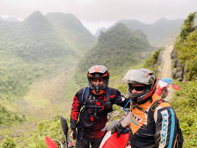 Two motorcyclists with helmets posing with lush mountainous landscape.