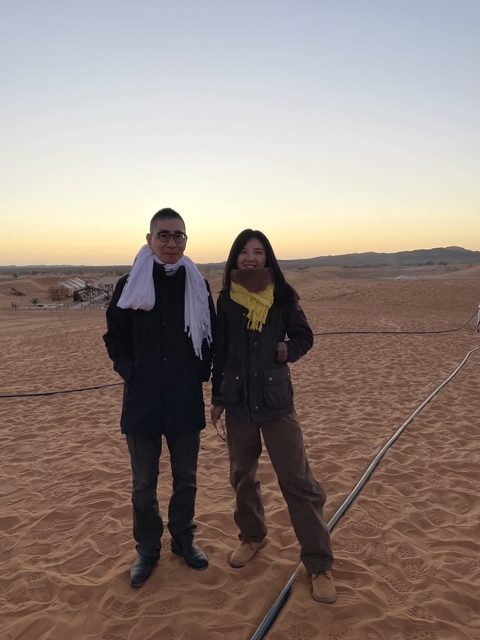       Two people standing in a desert landscape as the sun sets.
  