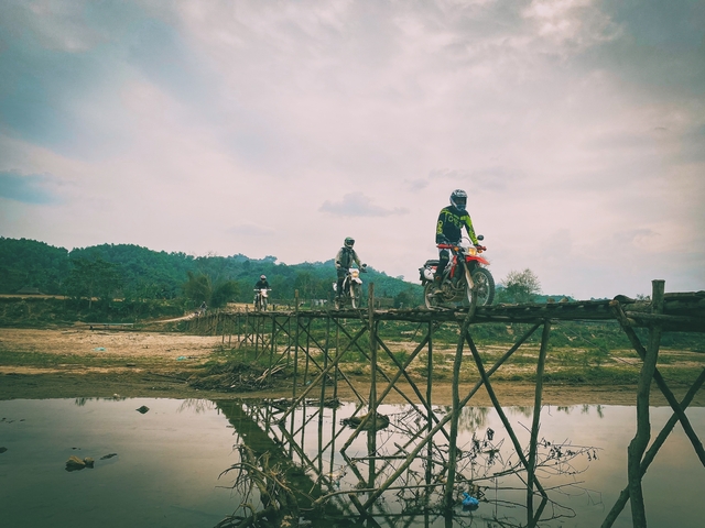 Motorbikes crossing a rustic wooden bridge over a reflective water body.