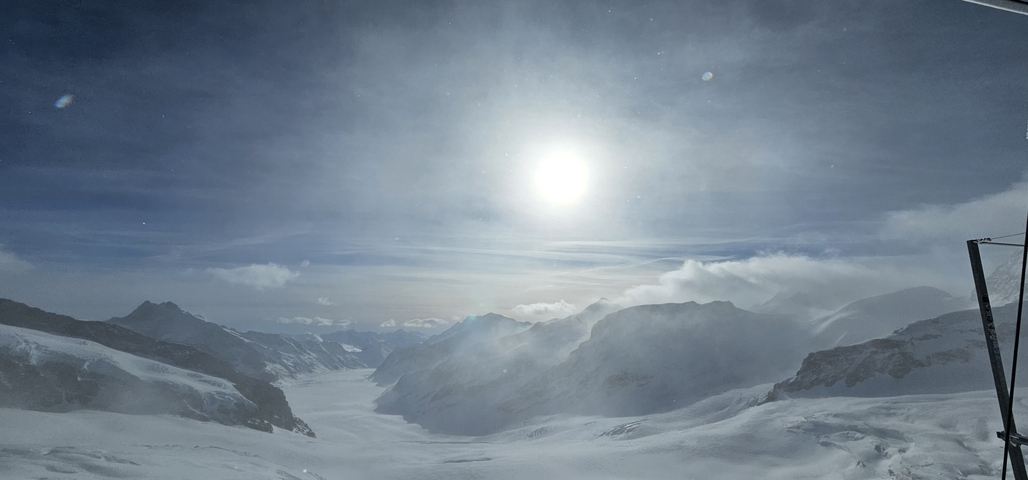       A snowy mountain landscape under a bright sun with clouds.
  