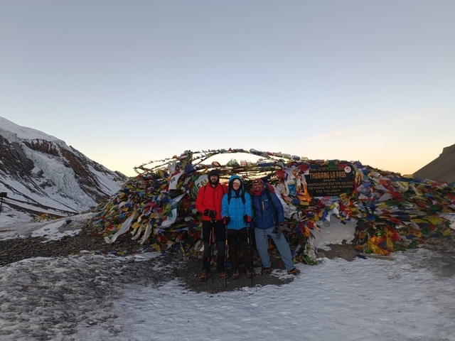 Three people posing with hiking sticks in front of a decorated pass with snow and prayer flags.