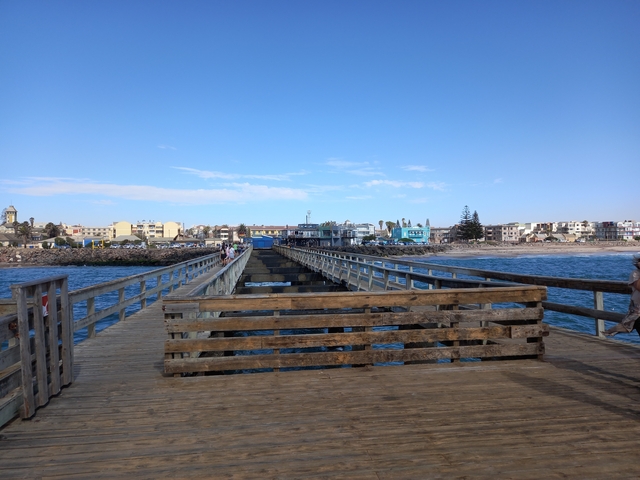 A wooden pier leading to the ocean with people walking on it and a town in the background.