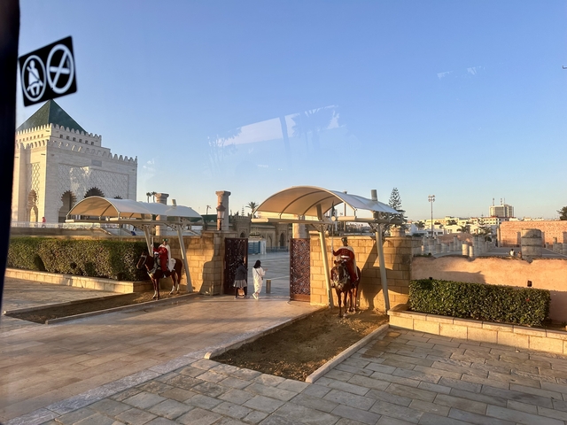       A gated entrance with guards on horseback, people nearby, and a building with a distinct rooftop.
  