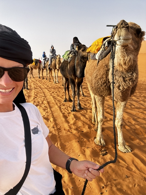       A person smiling with camels lined in the desert.
  