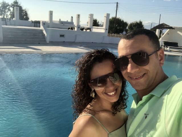       A couple posing near a swimming pool with white buildings in the background.
  