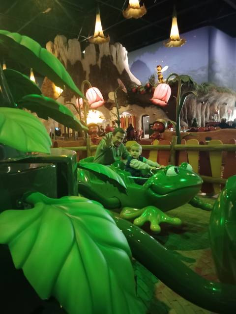 Adult and child on a green frog-themed amusement ride indoors.