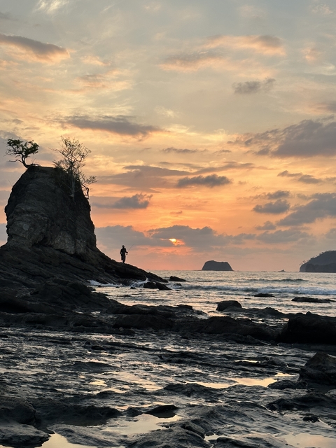       Person standing on a rock during a dramatic sunset over the ocean.
  