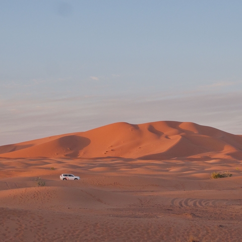       White SUV crossing a desert with sand dunes at sunset.
  
