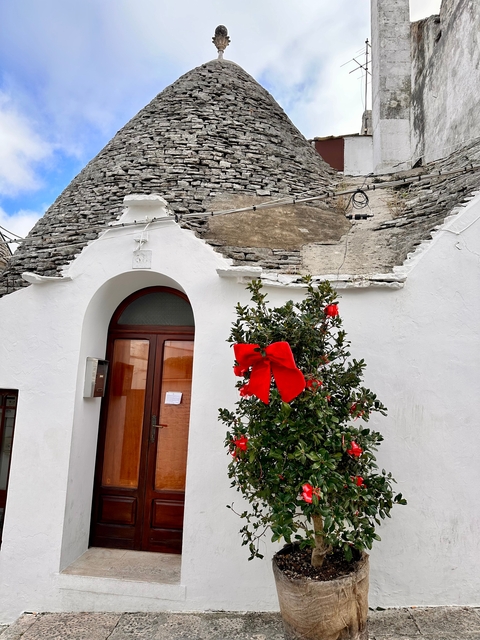 Traditional white conical building, possibly a trullo, decorated with a red bow.