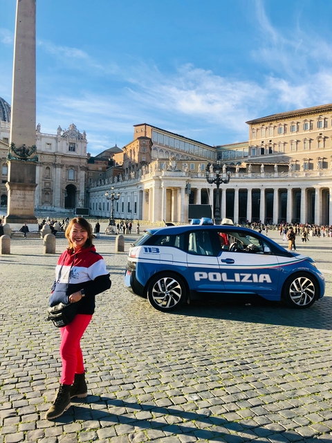      A woman smiling near a police car in a city square with historical architecture.
  