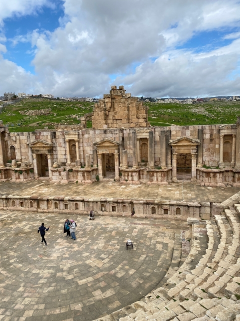 Ancient Roman amphitheatre with green hills and ruins in the background.
