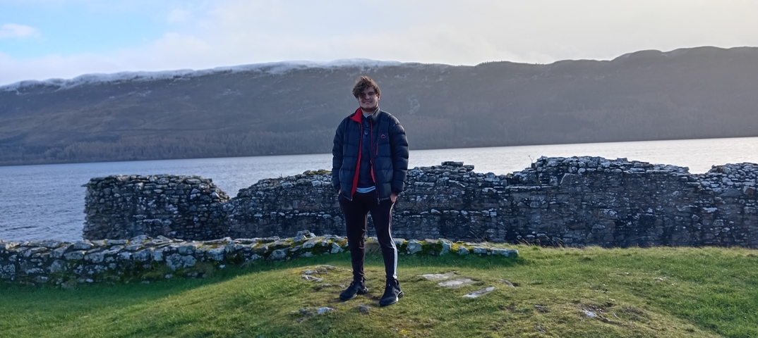       A person standing in front of ruins by a lake with hills in the background.
  