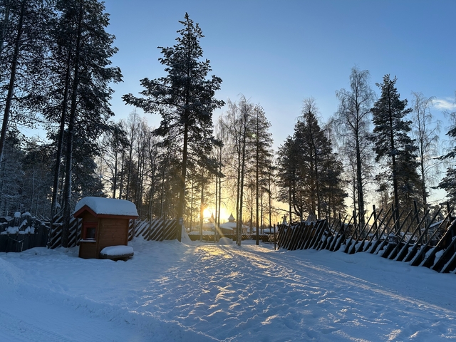       Snowy path with trees and a wooden structure, sun setting.
  