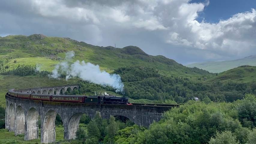 Steam train crossing a viaduct in a hilly, green landscape.