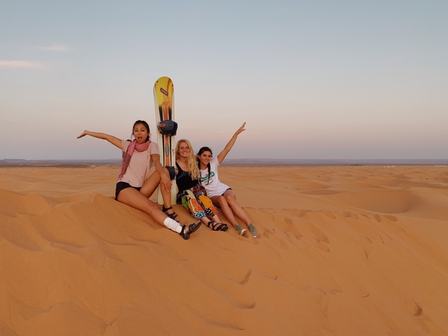       Three people sitting on sand dunes with a sandboard.
  