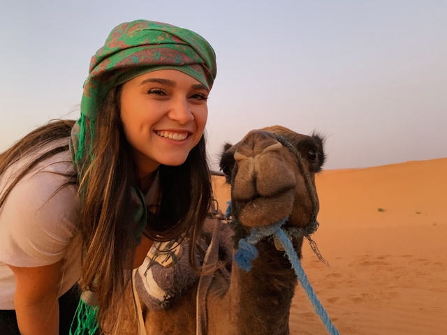       A smiling person with a camel in the desert.
  