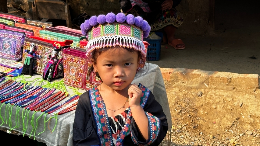       Child in traditional attire standing by a display of colorful textiles.
  