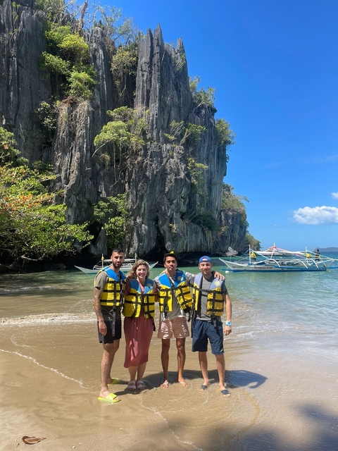 Four people in life jackets standing on a beach with boats and cliffs in the background.