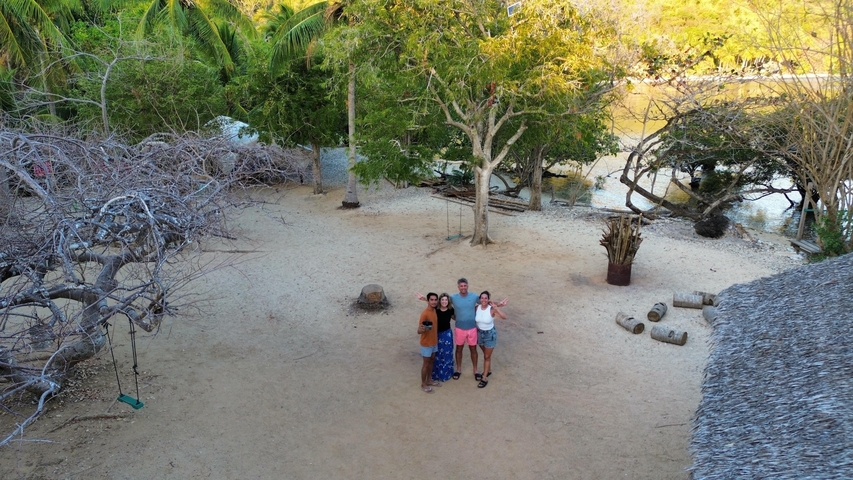 Group of people on a beach with trees and a thatched hut.