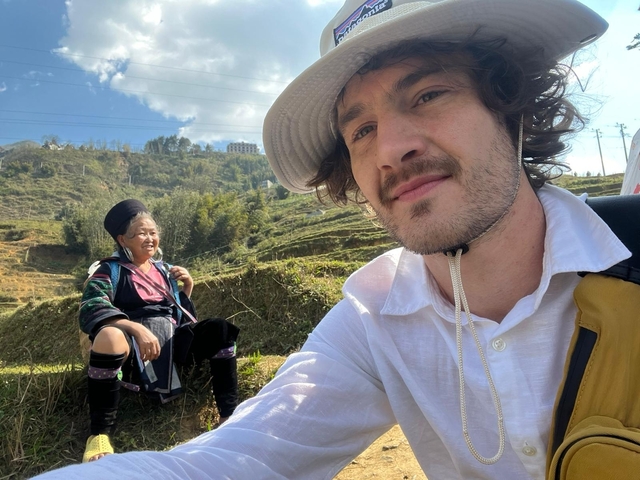       A close-up selfie with a backdrop of terraced fields and a local woman.
  