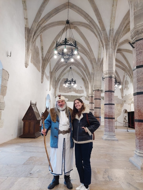       Two people standing indoors with a vaulted ceiling and chandeliers.
  
