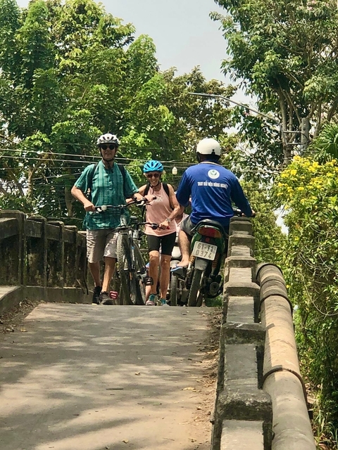       People cycling with a motorcycle on a small bridge.
  