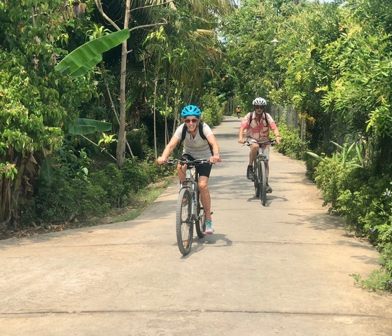Two people cycling on a road lined with green trees.