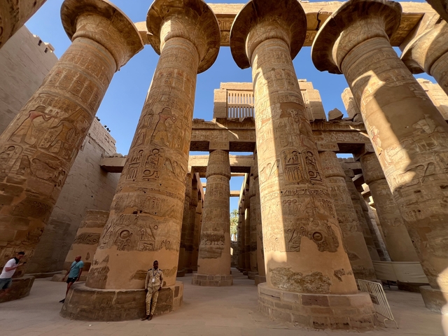       Inside a temple with carved columns and tourists walking.
  