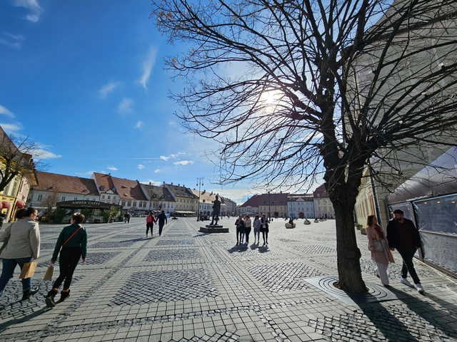       Open town square with cobblestone pathways, people walking, and cafes along the edge.
  