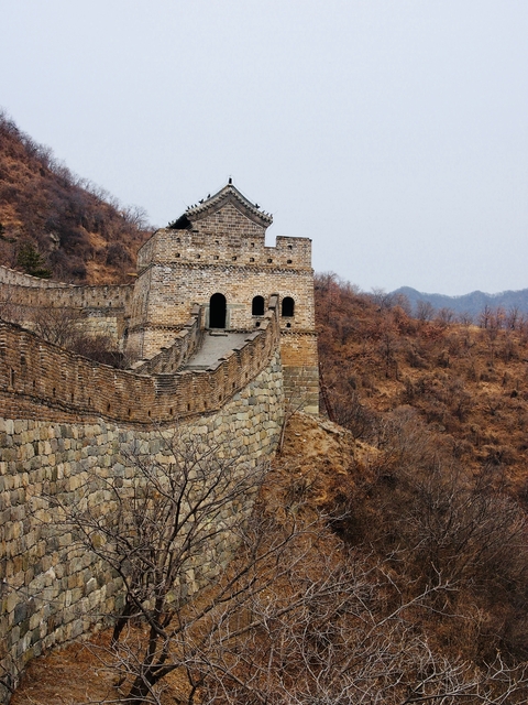       Stone watchtower along the Great Wall of China with bare trees surrounding it.
  