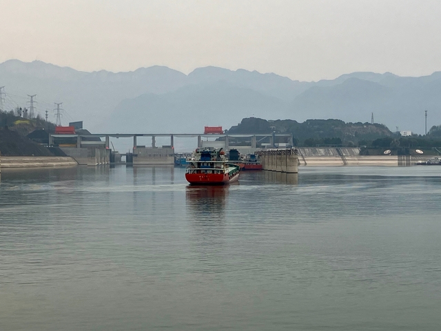 Cargo ships on a river with a dam and mountains in the distance.