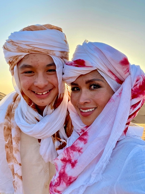       Two people smiling, wearing traditional headscarves in a desert setting.
  