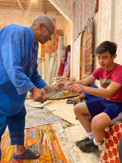       People enjoying tea in a carpet shop surrounded by rugs.
  