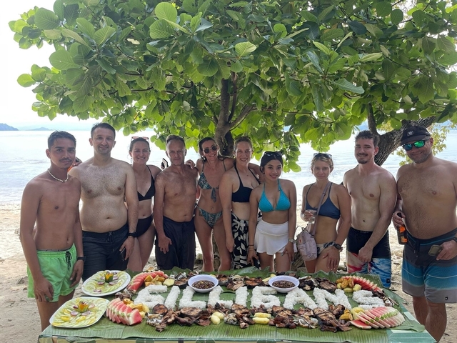       Group of people enjoying a meal on a beach under trees.
  