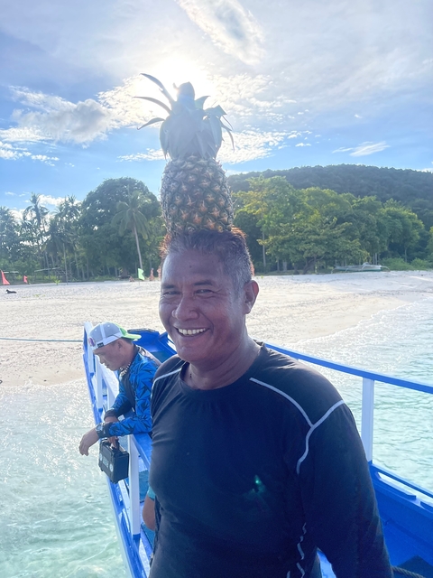       Man with a pineapple on his head smiling on a beach.
  