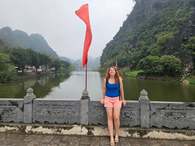       Person standing on a stone bridge with flag poles beside a calm river and mountains in the background.
  