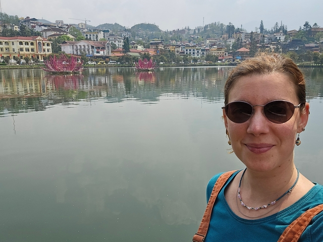       Person with sunglasses near a calm body of water with decorative boats and town in the background.
  