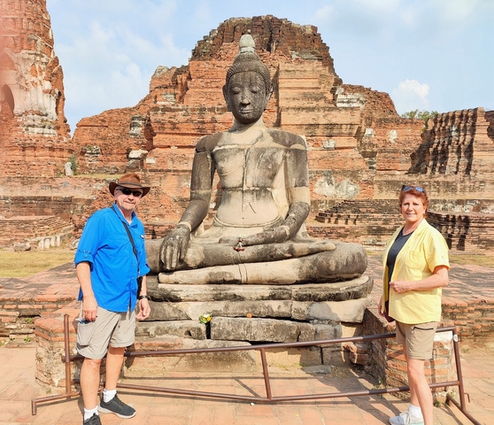       Two people standing beside a large Buddha statue in ruins.
  