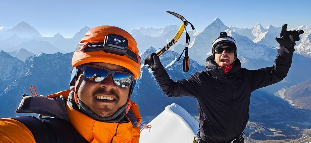 Two people in winter gear with ice axes on a snowy mountain with other mountains in the background.