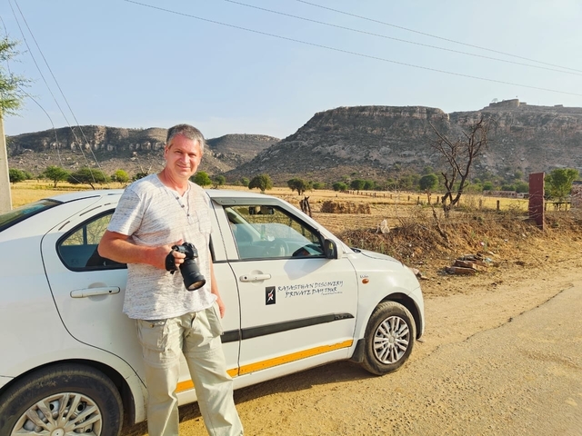 Man posing with camera by a car near hills.