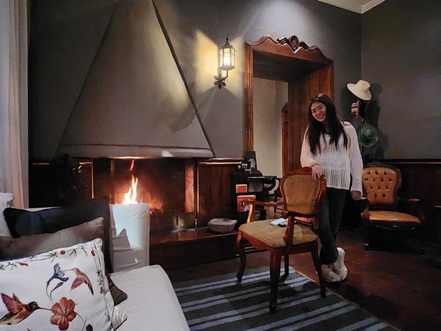 Person posing in front of a fireplace inside a cozy room.