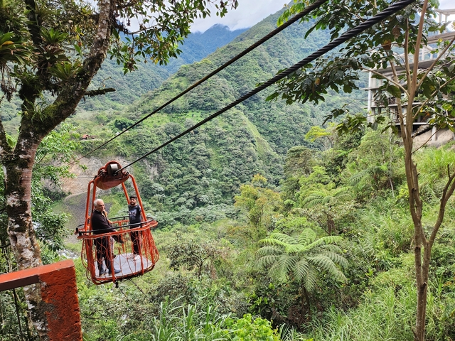       Two people in a cable basket crossing above a lush, mountainous landscape
  