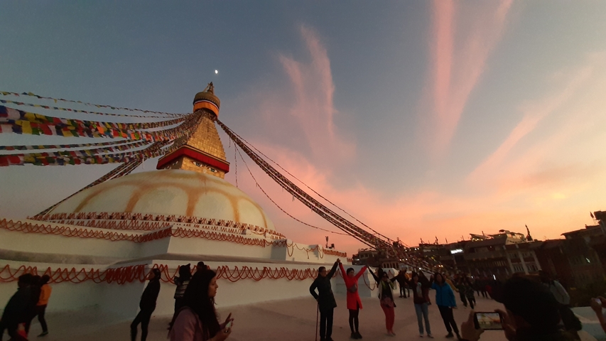       A large stupa with prayer flags during sunset, people around.
  