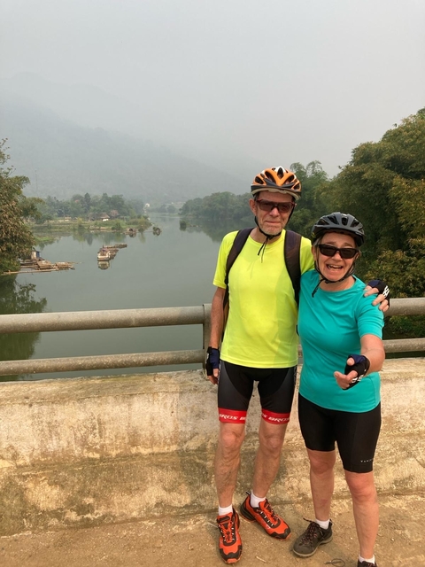 Two cyclists posing on a bridge over a river with a scenic backdrop.